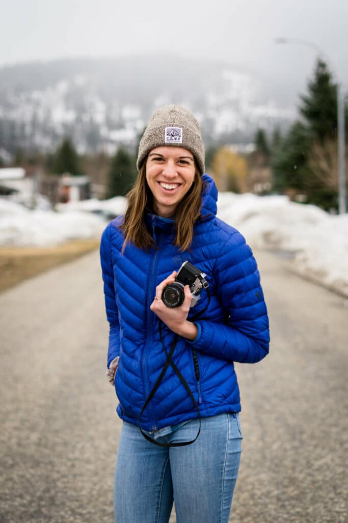 Smiling woman wearing toque and blue puffer jacket holding camera standing on road