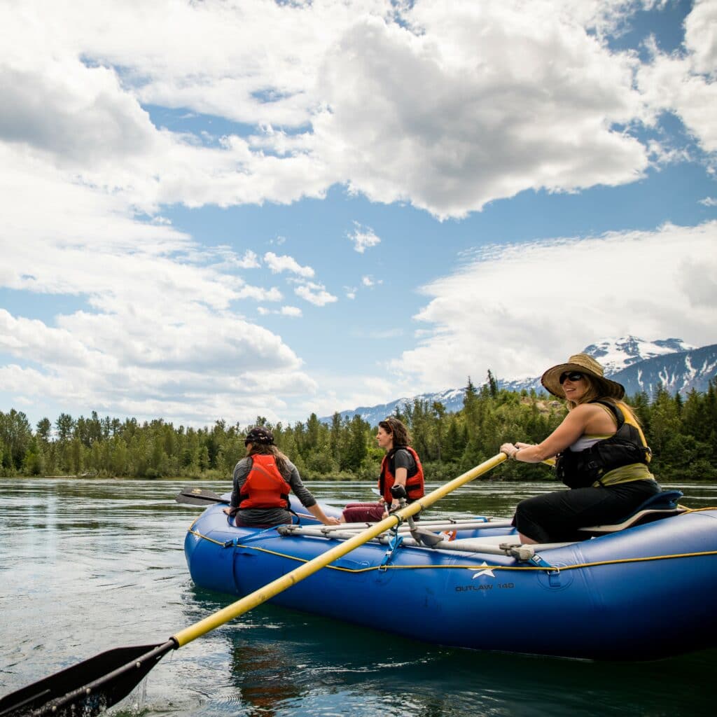 Three women in a blue raft paddling along a river with mountains and trees in the background