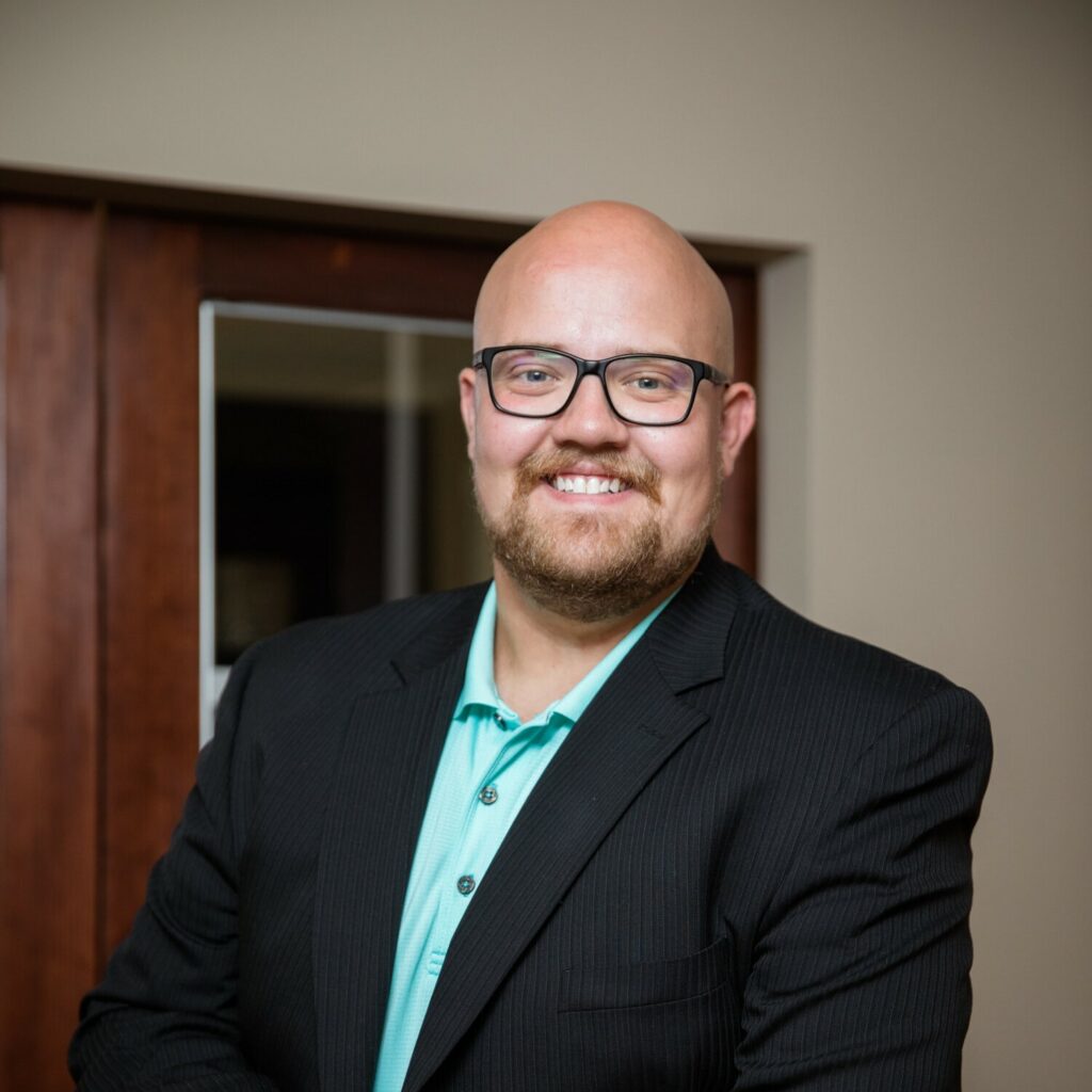 Smiling bald man with groomed beard wearing black rim glasses in a dark pinstripe suit and aqua shirt