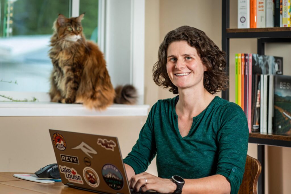 Smiling woman with brown curly hair sitting at laptop with cat in the background