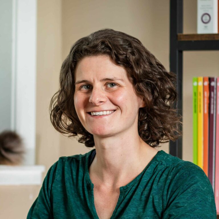 Smiling woman with brown curly hair wearing teal coloured top sitting in front of book shelf