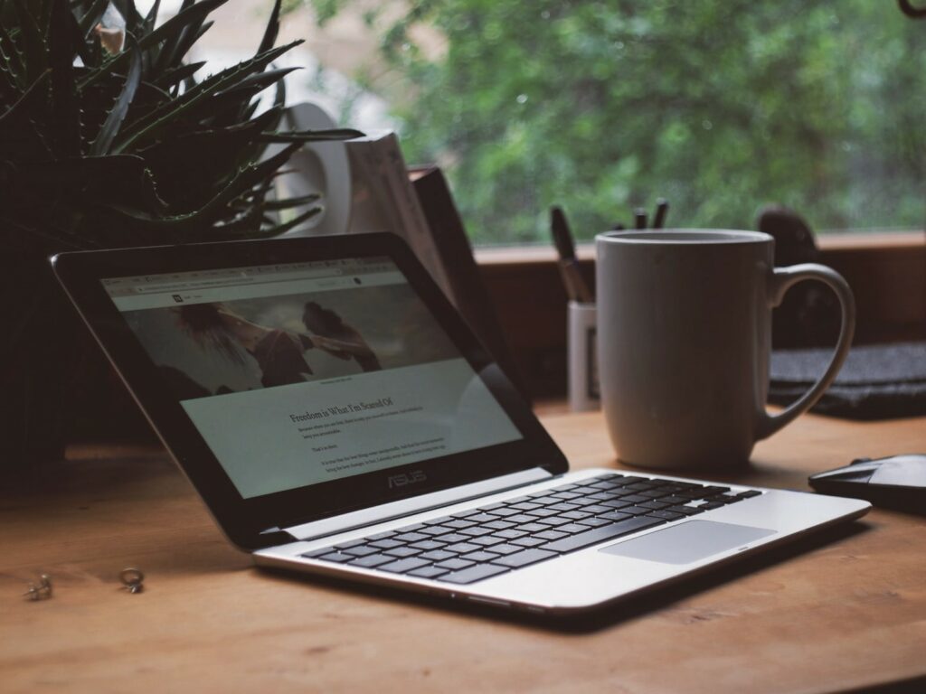 Open laptop sits on a wooden desk next to a white coffee mug and indoor plant with pens in the background